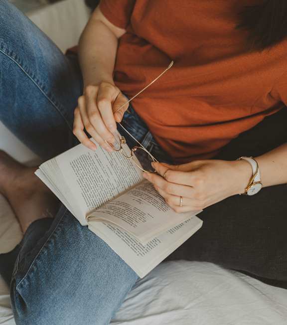 A person in an orange shirt is sitting on a bed and reading a book.
