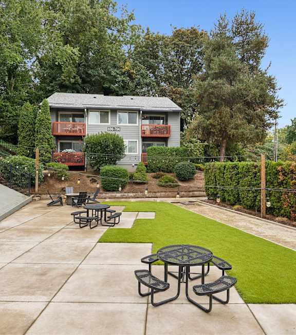 A patio with a table and chairs is surrounded by a green lawn and trees.