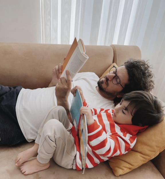 a father and son laying on a couch reading a book