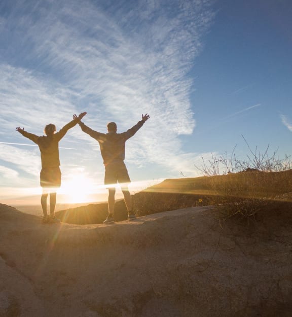 two people standing on a sand hill with their hands up in the air at sunset