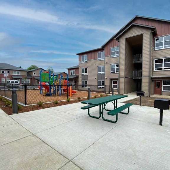 a playground with a slide and swings is in front of a building with a green picnic table in front of it.