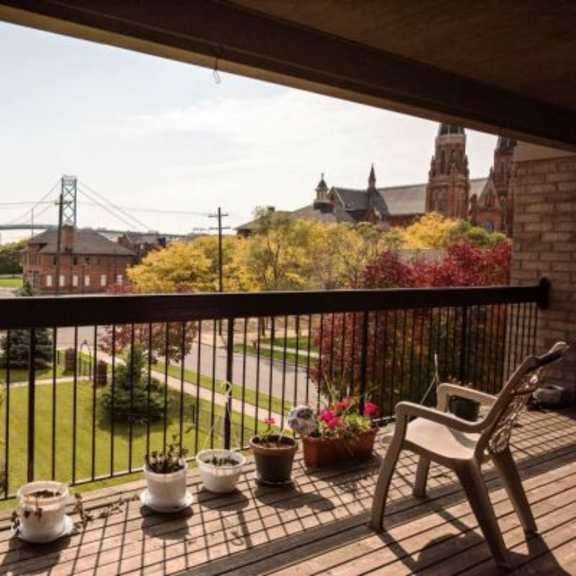 a balcony with a chair and potted plants overlooks a green lawn and a bridge.