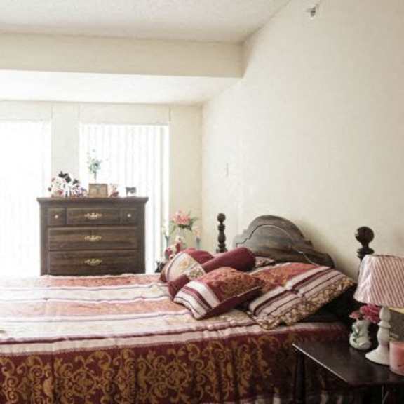 a bed with a red and gold comforter sits in a room with a wooden nightstand and a window.