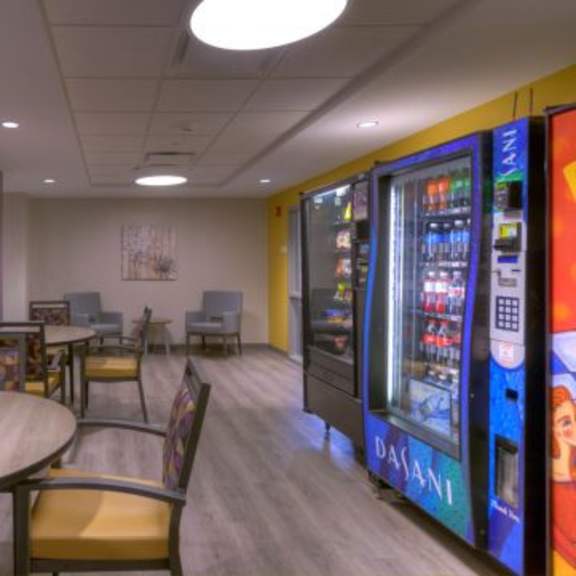 a room with a round table and chairs, a vending machine, and a wall with a picture.
