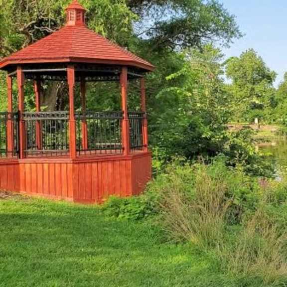 a red gazebo sits by a pond in a grassy field.