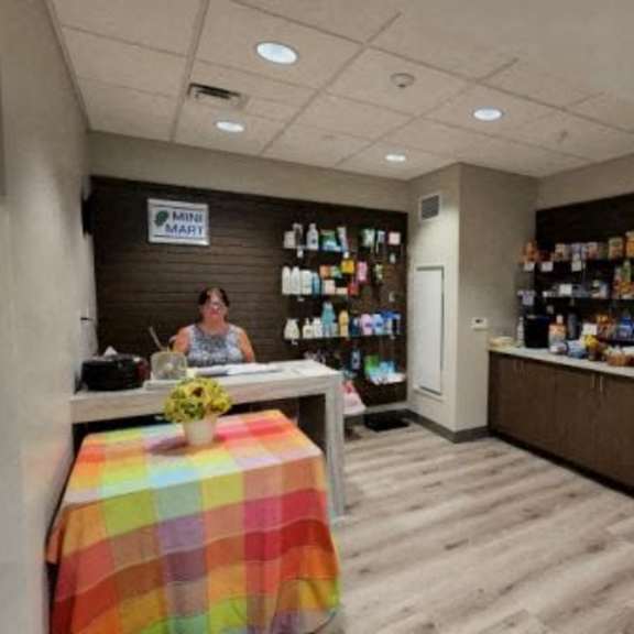 a woman is sitting at a desk in a room with a wall of products and a fridge.