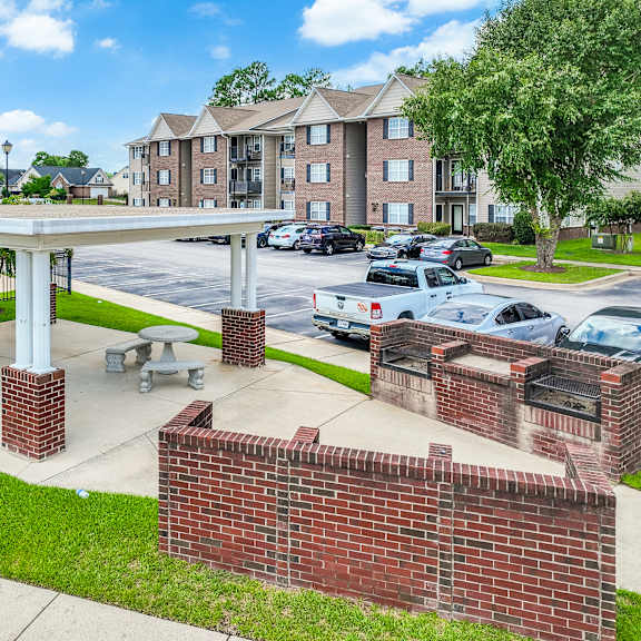 a covered outdoor picnic area with tables and benches is in front of a brick wall and apartment buildings.