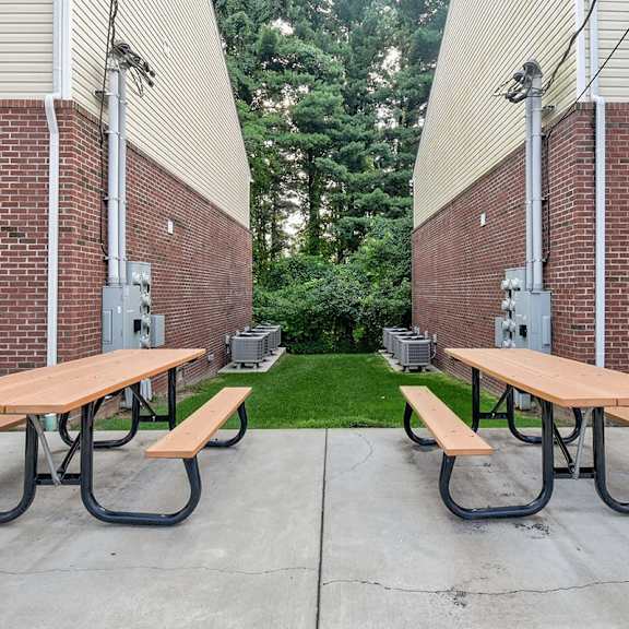 two picnic tables are set up on a concrete patio between two brick buildings.