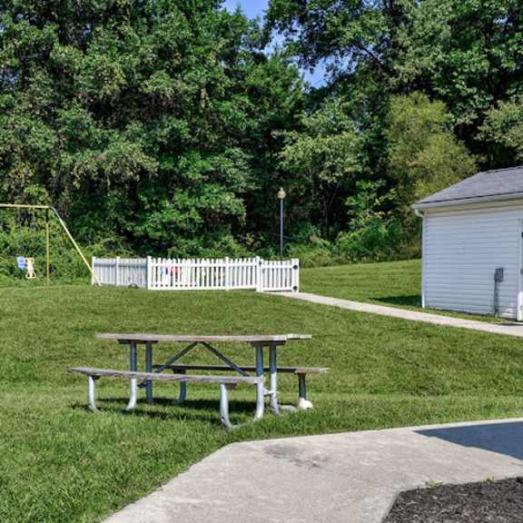 a picnic table is in the middle of a grassy area with a swing set and a white fence in the background.