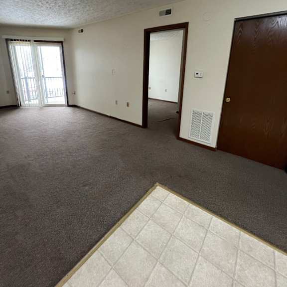 a room with a carpeted floor, a window with blinds, a door, and a wall-mounted air conditioning unit.