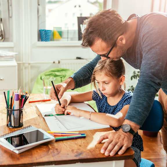 a father helping his daughter draw on a piece of paper at a desk