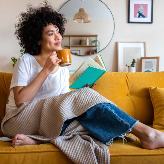 a woman with curly hair is sitting on a yellow couch, holding a mug and reading a book.