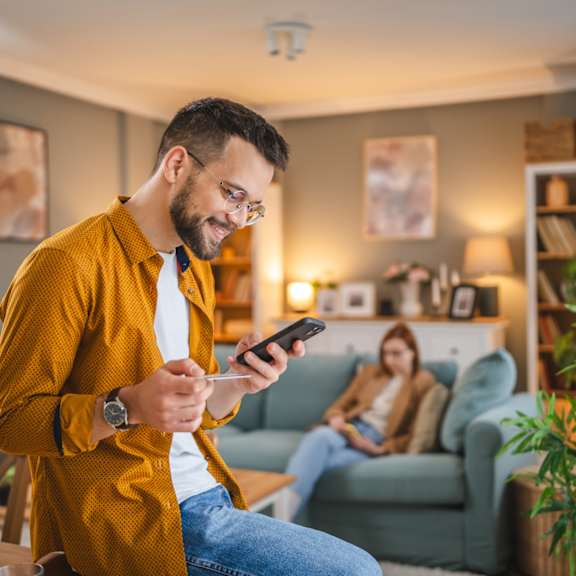 a man in a yellow shirt is looking at his phone while a woman sits on the couch in the background.