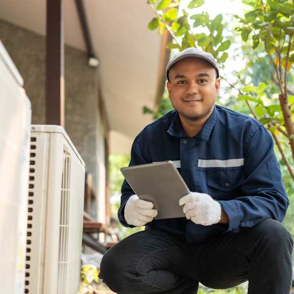 a man in a blue uniform is squatting by a white air conditioning unit, holding a clipboard.