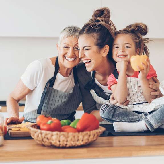 a woman and a young girl are laughing while sitting at a kitchen counter with a basket of vegetables and a bowl of salad in front of them.