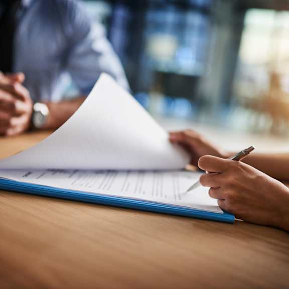 a person is writing on a notepad with a pen while another person is sitting across the table.