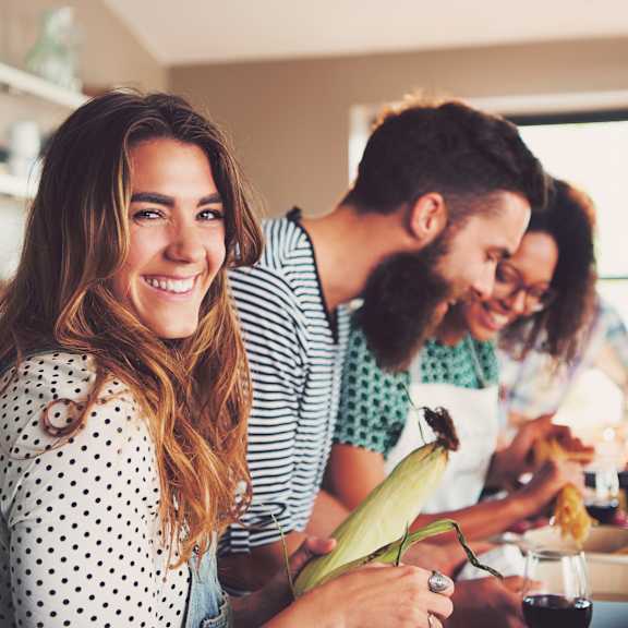 a woman with a polka dot shirt is holding a corn cob while two people prepare food in the background.