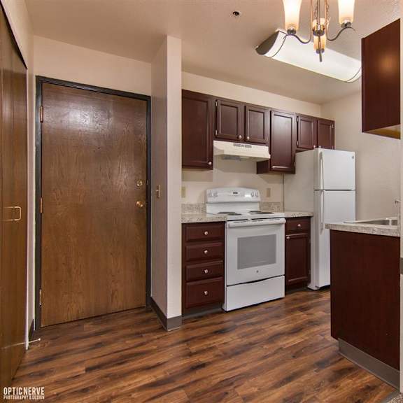 compact kitchen here at outlook apartments with dark wood cabinets, white appliances, and marble countertops. a wooden door and elegant light fixture add warmth and character.