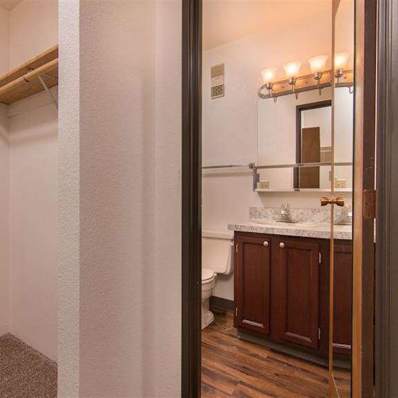 compact bathroom here at outlook apartments with wooden cabinets, marble countertop, and lit wall mirror seen from a doorway. adjacent closet with a wooden shelf. warm and clean ambiance.