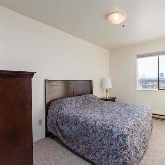 simple bedroom here at outlook apartments with a paisley-patterned bed, wooden headboard, and nightstand with lamp. window shows cityscape, creating a serene atmosphere.