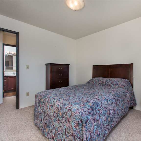 a cozy bedroom here at outlook apartments features a bed with a colorful paisley bedspread, a wooden nightstand with a brass lamp, a tall dresser, and a doorway leading to a bathroom.