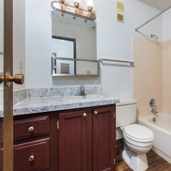 compact bathroom here at outlook apartments with granite countertop, wooden cabinets, and a wall-mounted mirror. beige tile surrounds the bathtub and shower, creating a clean, simple look.