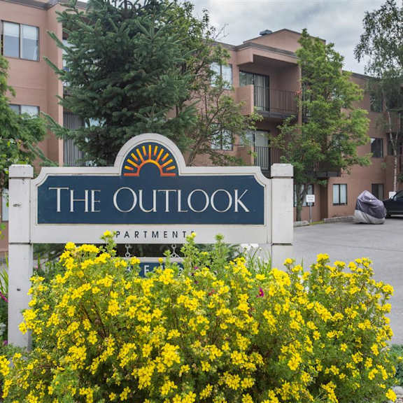 sign reading "the outlook apartments" with a sun icon. surrounded by vibrant yellow flowers, trees, and a brown apartment building in the background. cozy atmosphere.