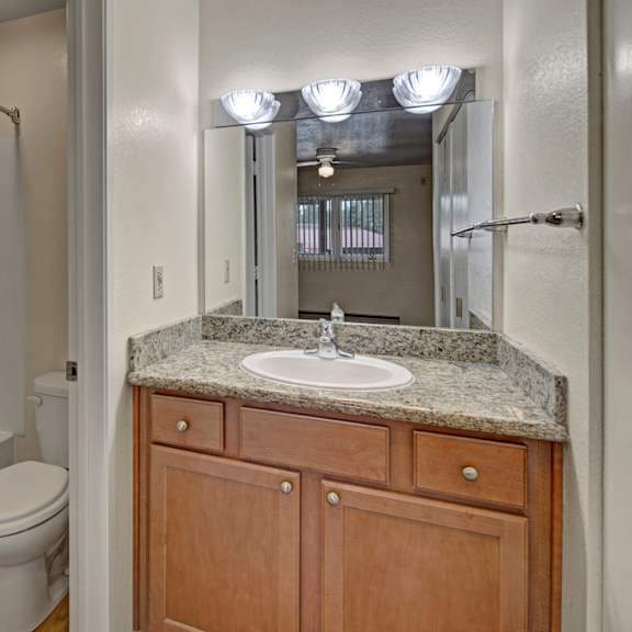 a clean and functional bathroom here at club at eagle pointe featuring a granite-style vanity countertop with a single sink, warm wood cabinetry, and a large wall mirror with bright overhead lighting. the space includes a tub and shower combination with built-in shelving, durable flooring, and a layout designed for everyday comfort and convenience.