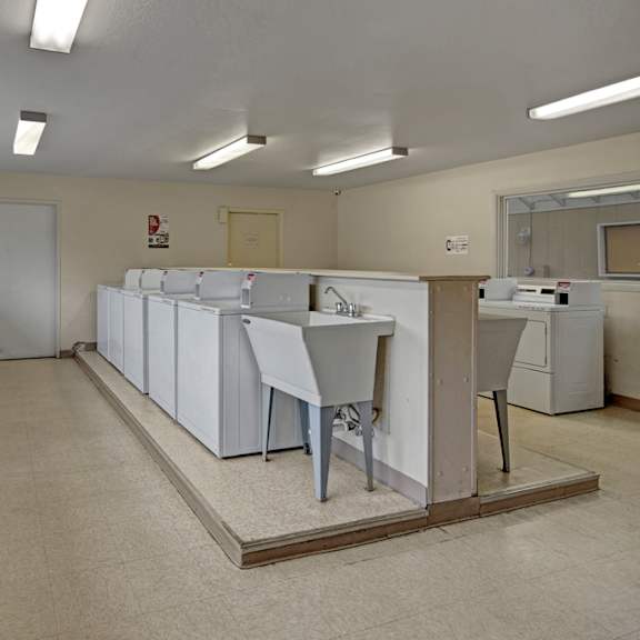 a clean, well-lit on-site laundry room here at club at eagle pointe featuring multiple washers and dryers, a utility sink, wide walkways, bright overhead lighting, and durable flooring, providing residents with a convenient, functional space for everyday laundry needs.