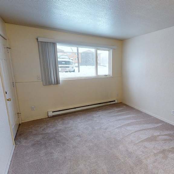 a spacious, sunlit bedroom here at club at eagle pointe featuring soft neutral walls, plush carpet flooring, and a wide window with vertical blinds that bring in natural light. a baseboard heater runs beneath the window, and the simple rectangular layout offers ample space for a bed, dresser, and additional furnishings.