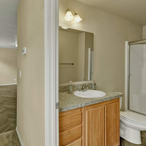 a clean, neutral bathroom here at four seasons featuring a granite-style vanity with wood cabinetry, a single sink, and an overhead mirror with warm lighting. a tub and shower combination with sliding glass doors sits beside the toilet, while the adjacent hallway showcases wood-style flooring that connects smoothly to the rest of the apartment.