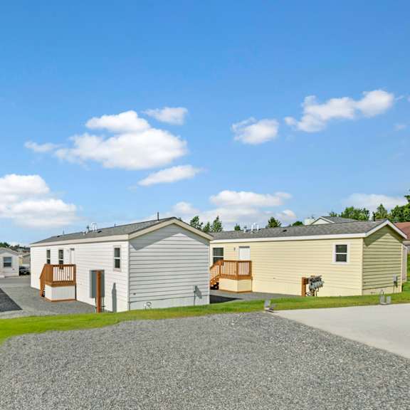 a peaceful community view here at four seasons showcasing well-maintained homes with neutral exterior siding, private entry decks, and neatly arranged pathways. bright blue skies, open green space, and a clean gravel drive create a welcoming neighborhood atmosphere that highlights the spacious layout and residential charm of the community.