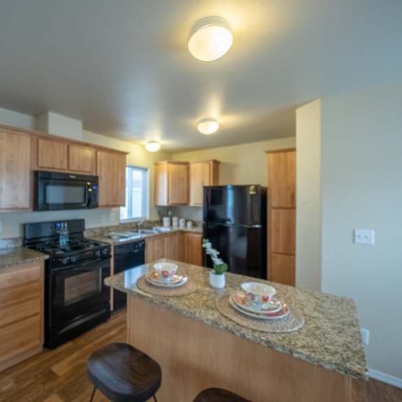 a bright, modern kitchen here at four seasons showcasing natural wood cabinetry, granite-style countertops, and a spacious center island with seating, paired with black appliances, warm wood-look flooring, ceiling lighting, and an open layout that offers ample prep space and an inviting flow into the surrounding living area.