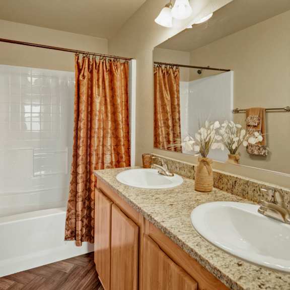 a spacious bathroom here at four seasons featuring a double-sink vanity with a granite-style countertop, light wood cabinetry, wide wall mirror with overhead lighting, a clean white tub and shower surround with a warm-toned curtain, neutral walls, coordinated towels, and wood-style flooring creating a comfortable, inviting space.