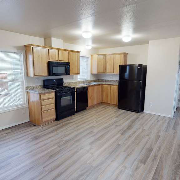 a bright open kitchen here at four seasons featuring light wood cabinetry, granite-style countertops, and black appliances that create a clean, modern look. wide plank flooring flows throughout the space, while large windows and ceiling lights provide ample natural and overhead lighting, connecting seamlessly to the adjacent living area and hallway.