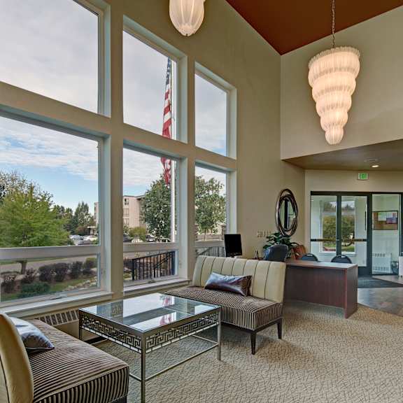 a bright lobby at the alpine with tall windows letting in natural light and views of the community. stylish seating surrounds a glass-top coffee table, and a desk sits near the entrance. modern lighting, warm carpeting, wall art, and indoor plants create an inviting welcome area.