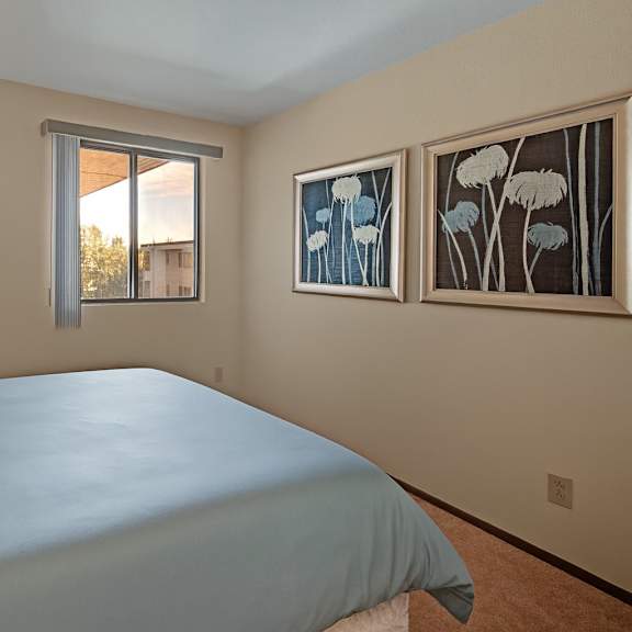 a peaceful bedroom at the alpine with a neatly made bed in soft blue bedding and patterned pillows. a window with vertical blinds lets in natural light and offers outdoor views. framed floral artwork decorates the wall, adding a calm and stylish touch to the space.