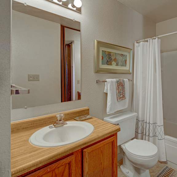 a clean bathroom at the alpine featuring a bathtub and shower combo with a white curtain. the vanity includes a single sink, wood cabinetry, and a large wall mirror with bright lighting. neutral walls, framed artwork, and simple towels add a warm, inviting touch.