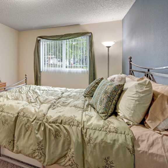 cozy bedroom here at brighton wood apartments with a green bedspread and matching pillows, a wooden dresser with decorative items, and a window with green curtains, creating a serene vibe.