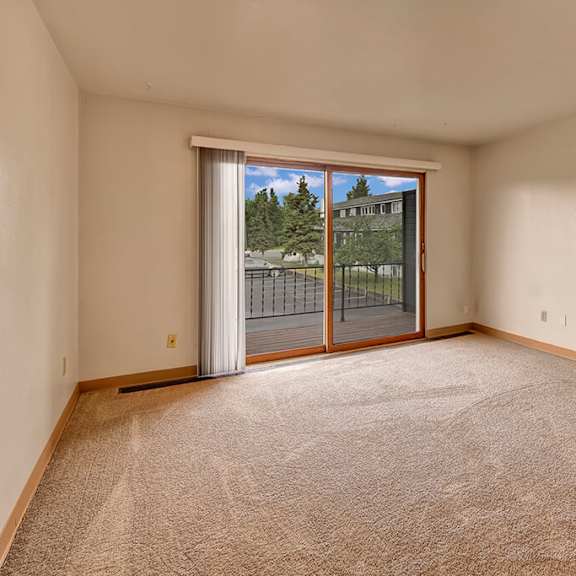 an empty living room with beige carpet and white walls. a large sliding glass door opens to a balcony with a view of trees and buildings, conveying a serene atmosphere. the alaskan apartments.