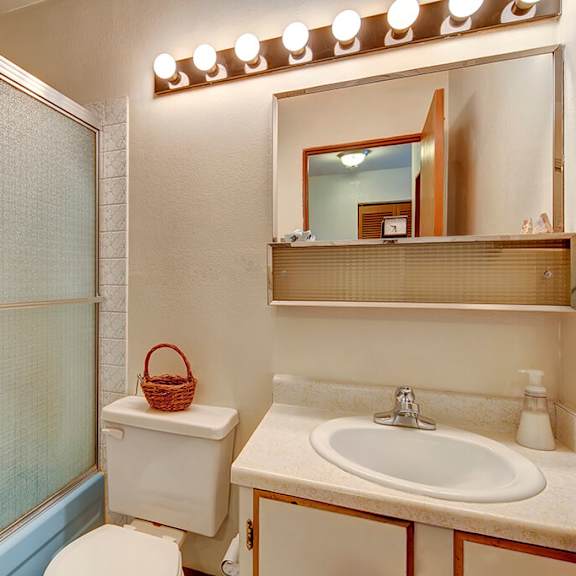 compact bathroom with a glass shower door, white toilet, and beige countertop with a sink. a wicker basket is on the toilet tank and bright lights above a mirror. the alaskan apartments.