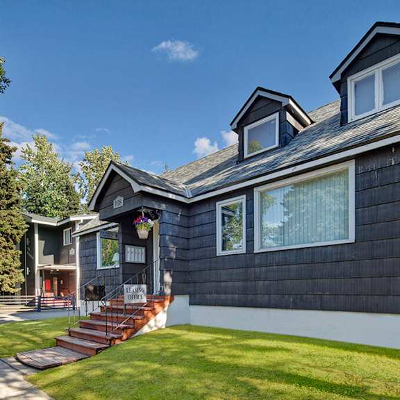 a quaint black house with white trim under a clear blue sky, featuring dormer windows and a flower pot. the sign reads “leasing office,” evoking a welcoming vibe. the alaskan apartments.