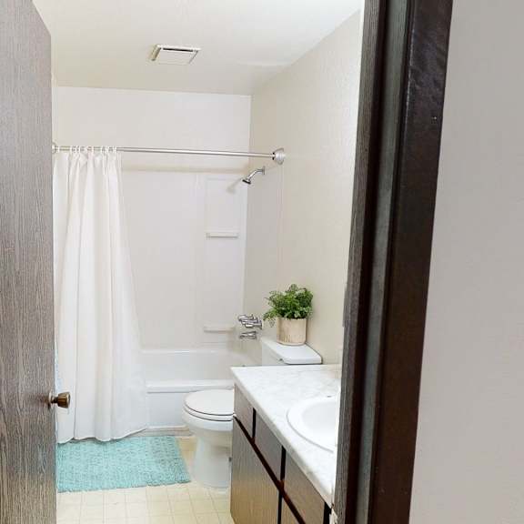 view from an open doorway into a small bathroom here at deer park apartments featuring a white shower curtain, a sink with a marble top, and a potted plant.