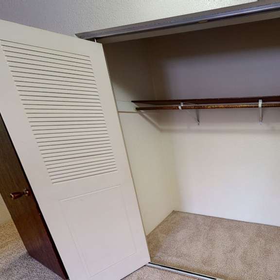 interior view of an empty closet here at deer park apartments with a carpeted floor, a brown shelf, and a hanging rod for clothes, flanked by paneled doors.