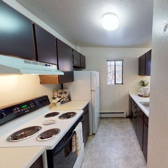 narrow kitchen here at deer park apartments with dark cabinets, white appliances, and tiled floor. bright overhead light, window, and decorative shelf enhance a cozy feel.