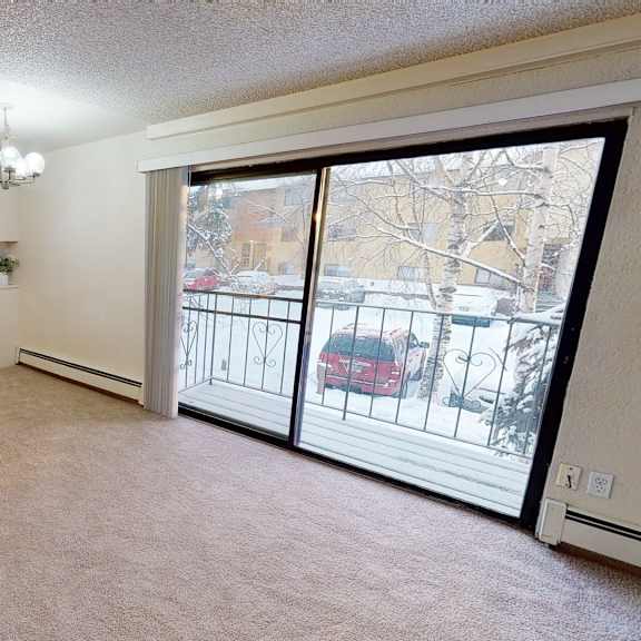 cozy apartment interior here at deer park apartments with beige carpet, a small kitchen with dark cabinets, and large sliding glass doors leading to a snowy balcony view.