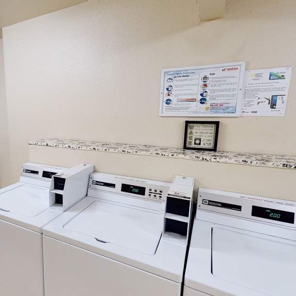 a clean laundry room here at hillside chalet apartments with stacked dryers on the left and top-loader washers on the right. instructions are mounted on a beige wall, creating an orderly atmosphere.