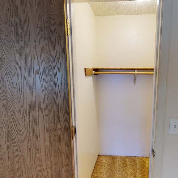 open closet here at rancho tudor apartments with wooden door ajar, revealing a white interior, a wooden hanging rod, and beige carpet. the space is minimal and tidy.