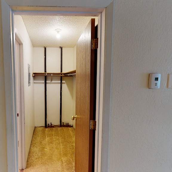 a small, empty closet here at rancho tudor apartments with beige walls and carpet, an open wooden door, a single light bulb, metal rods, wooden shelves, and a wall thermostat.