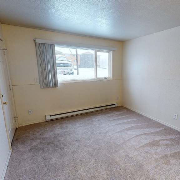empty living room here at the club at eagle pointe apartments with beige walls and carpet, featuring a large window with vertical blinds. sunlight highlights the carpet's texture, creating a calm atmosphere.